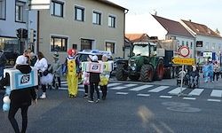 Menschen in Kostümen gehen eine Straße entlang. Einer hält eine Box mit der Nummer 45. Ein Traktor und ein Gebäude im Hintergrund.