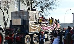 Ein dekorierter Lastwagen, möglicherweise für einen Umzug, mit Menschen, die darauf reiten. Viele Menschen beobachten ihn auf der Straße.