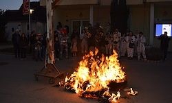 Ein brennendes Lagerfeuer brennt hell in der Nacht vor einem Gebäude. Eine Menschenmenge steht herum und beobachtet das Feuer. Einige halten brennende Fackeln.