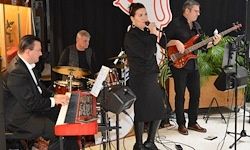Eine Band tritt auf der Bühne auf, mit einer Sängerin und einem Gitarristen. Ein anderer Mann spielt sitzend Klavier.