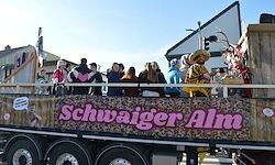 Ein großer dekorierter Lastwagen mit Menschen obenauf, darunter eine Person mit einem Sombrero. Der Lastwagen hat ein Schild, das Schwaiger Alm besagt.