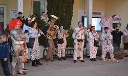 Eine Gruppe von Menschen in Kostümen mit Musikinstrumenten, wahrscheinlich bei einer Straßenshow. Einige sind in Clown-Kostümen, während andere in Tupfen-Outfits sind. Einige spielen Blechblasinstrumente.