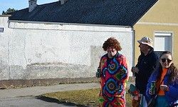 Drei Menschen gehen auf dem Bürgersteig, einer in farbenfroher Kleidung und zwei weitere mit Hüten. Ein Gebäude mit gelben Wänden steht hinter ihnen.
