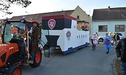 Ein Umzug mit einem Traktor, der einen geschmückten Wagen zieht, eine Person in Kostüm darauf und mehrere Menschen in Kostümen dahinter.