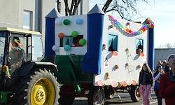 Ein Traktor mit einem geschmückten Anhänger in Blau und Weiß fährt mit Menschen um ihn herum die Straße entlang.