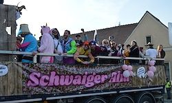 Ein mit Menschen in Kostümen gefüllter Wagen, der 'Schwaiger Alt' beschriftet ist. Die Menschen winken und halten Luftballons. Der Wagen ist im rustikalen Stil mit Holzakzenten dekoriert.