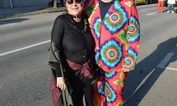 Zwei Personen in leuchtenden Outfits stehen auf einer Stadtstraße, wobei einer ein farbenfrohes Tie-Dye-Kostüm trägt und der andere in einem dunklen Outfit mit einer grünen Jacke. Sie sind von Stadtelementen wie Straßenmarkierungen und Schatten umgeben.