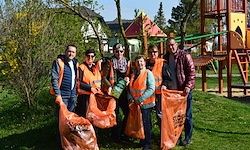 Eine Gruppe von Menschen in orangenen Westen steht auf einem Rasen, hält orangene Taschen und posiert für ein Foto in der Nähe eines Spielplatzes.