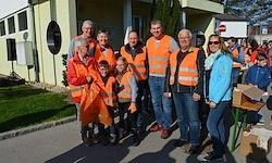 Eine Gruppe von Menschen in orangewesten steht vor einem gelben Gebäude. Sie posieren für ein Foto mit einem Rasenbereich, einem Tisch mit Kisten und einer Menschenmenge im Hintergrund.