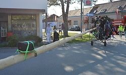 Ein Mann steht vor einem langen Holzbalken mit einem Lastwagen im Hintergrund. Menschen gehen auf der Straße.
