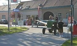 Ein grüner Traktor ist auf der Straße geparkt. Menschen stehen um ihn herum, einige tragen Shorts. Im Hintergrund sind Gebäude, Autos und Bäume zu sehen.