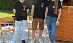 Drei Personen stehen in einem Garten zusammen, mit einer Holzkonstruktion hinter ihnen. Zwei Männer und eine Frau tragen schwarze T-Shirts. Der Mann auf der linken Seite hat einen Fleck auf seinem Hemd.