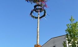 Ein hoher Pfahl mit zwei Kränzen steht vor einem Gebäude bei klarem blauem Himmel im Hintergrund.