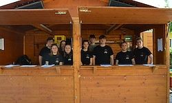 Eine Gruppe von Menschen in schwarzen T-Shirts steht in einer Holzkabine, lächelt und posiert für ein Foto.