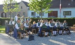 Eine Brassband spielt auf einer Terrasse mit Grünpflanzen, Gebäuden und einer Flagge im Hintergrund.