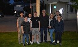 Eine Gruppe von Menschen steht auf einem Rasen vor einem Gebäude, das bei Nacht von Straßenlaternen beleuchtet wird. Sie posieren für ein Foto.