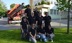 Eine Gruppe von Menschen in schwarzen T-Shirts und Sonnenbrillen posiert für ein Foto auf einem Rasen, mit einem Feuerwehrwagen und einem Gebäude im Hintergrund.