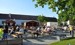 Ein Biergarten mit mehreren Tischen und Bänken, viele Menschen sitzen und stehen, Bäume und ein Haus im Hintergrund.