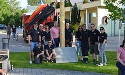 Eine Gruppe von Menschen in Schwarz posiert für ein Foto vor einem Denkmal mit einem roten Kran im Hintergrund. Ein Gebäude ist hinter ihnen zu sehen.