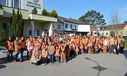 Eine große Gruppe von Menschen in orangefarbenen Westen posiert für ein Foto vor einem Gebäude mit dem Schild 'Amtsstrasse Schwadorf'.