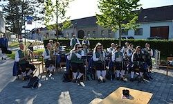 Eine Musikergruppe spielt Instrumente auf einer gepflasterten Straße vor einem Gebäude. Einige Leute sitzen, andere stehen. Der Bereich ist von Bäumen und Büschen umgeben.
