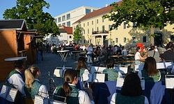 Ein Musikensemble in grünen Westen und weißen Hemden tritt auf einer Freiluftbühne auf einem Stadtplatz mit Gebäuden und Menschen auf.