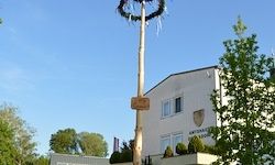 Ein hoher Pfahl mit einem Kranz und einem Schild steht vor einem weißen Gebäude mit vielen Fenstern und einem Balkon, vor dem Hintergrund von Bäumen und blauem Himmel.