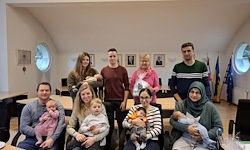 Eine Gruppe von Erwachsenen und Kindern, darunter einige mit Babys, posiert für ein Foto in einem Raum mit gewölbten Fenstern, Flaggen und Bilderrahmen an der Wand.
