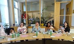 Eine Gruppe von Frauen und Kindern sitzt um einen Tisch mit Flaschen und Tellern. Einige halten Babys. Sie sind in einem Raum mit einer Treppe und Pflanzen.