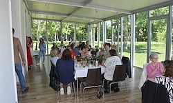Eine Gruppe von Menschen sitzt an Tischen in einem glaswandigen Raum, wobei eine Person in der Nähe geht. Der Raum hat Holzböden und Blick auf einen Rasenbereich mit Bäumen.