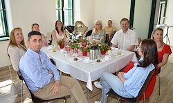 Eine Gruppe von Menschen sitzt um einen Tisch in einem Raum. Sie lächeln und scheinen für ein Foto zu posieren. Der Tisch hat Blumen, eine silberne Karaffe und Tassen. Der Raum hat einen Holzboden und weiße Wände mit grünen Fensterrahmen.