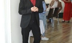 Ein Mann in einem Anzug steht an einer weißen Wand und schaut nach unten. Hinter ihm sitzen zwei Personen an einem Tisch, und eine weitere steht in einem roten Kleid.
