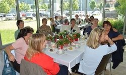 Eine Gruppe von Frauen sitzt um einen Tisch mit Blumen und Tellern in einem Restaurant. Sie essen. Draußen sind Autos geparkt.