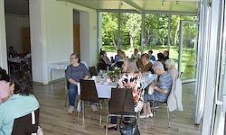 Eine Gruppe von Menschen sitzt um Tische in einem großen Raum mit Glaswänden, mit Blick auf eine üppige grüne Landschaft.