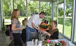 Ein Mann überreicht einer älteren Frau, die an einem Tisch in einem Restaurant sitzt, einen Blumenstrauß. Eine weitere Frau steht in der Nähe, und sie sind von einer Gartenansicht umgeben.