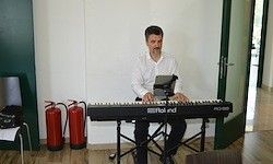 Ein Mann spielt Klavier in einem Raum mit weißen Wänden, Holzboden und zwei roten Feuerlöschern. Er trägt ein weißes Hemd und schwarze Hose.