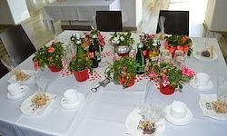 Ein Esstisch ist mit Tassen, Untertassen, Blumen und Flaschen gedeckt. Der Tisch ist mit einer weißen Tischdecke bedeckt.