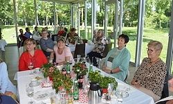 Eine Gruppe von Menschen sitzt um einen Tisch mit Blumen, Tassen und Flaschen. Der Tisch ist mit einer weißen Tischdecke bedeckt. Sie sitzen in einem Glasraum mit einem Grasfeld im Hintergrund.