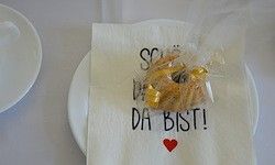 Ein Teller mit einer Serviettenkappe oben hält ein Gebäck in Plastikfolie, mit den Worten 'So da bist!' darauf geschrieben.