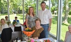 Eine Gruppe von Menschen steht in einem Gartenrestaurant. Eine Frau hält einen Blumenstrauß und sitzt neben einer anderen Frau. Sie stehen mit einem Mann und alle lächeln.