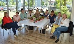 Eine Gruppe von Frauen sitzt um einen Tisch in einem Raum mit Glasfenstern, einem Holzboden und Pflanzen draußen. Ein paar Autos sind draußen geparkt.