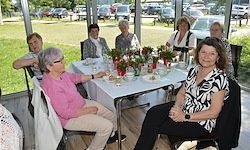 Eine Gruppe von Menschen sitzt um einen Tisch mit Blumen, Tellern und Tassen. Sie befinden sich in einem Restaurant mit großen Fenstern und einem Grasfeld draußen.