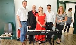 Fünf Personen stehen um ein digitales Klavier herum. Eine Frau in einem roten Kleid steht vor dem Klavier. Zwei Männer und zwei Frauen stehen hinter dem Klavier.