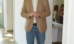 Ein Mann in einem beigen Blazer und blauer Jeans steht in einem Flur, Hände vor der Brust verschränkt, mit einem verschwommenen Hintergrund eines Raums mit Personen und einem Tisch.