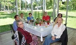 Eine Gruppe von Frauen sitzt um einen Tisch in einem Raum mit Glaswänden, umgeben von einer grünen Wiese. Der Tisch ist mit einer weißen Tischdecke bedeckt und hat Blumen, Tassen und Flaschen.