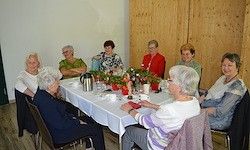 Eine Gruppe älterer Frauen sitzt um einen Tisch mit Blumen, Tassen und einem Krug. Sie sind in einem Raum mit Holzwänden und -böden. Einige tragen Brillen und Halsketten.
