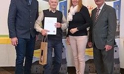 Vier Personen stehen in einer formellen Umgebung. Ein Mann hält ein Zertifikat und eine Tasche. Eine Frau hält seinen Arm. Dahinter ist ein Hintergrund mit Bildern.
