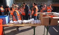 Eine Gruppe von Menschen in orangenen Westen steht um einen Tisch mit Kisten herum und bereitet Essen für eine Gemeinschaftsveranstaltung vor.