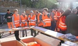 Eine Gruppe von Menschen in orangefarbenen Warnwesten steht um einen Tisch mit Kisten und Ausrüstung herum, möglicherweise Freiwillige, die an einer Gemeinschaftsveranstaltung oder einer Reinigung teilnehmen.
