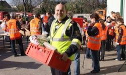 Ein Mann in einem gelben Sicherheitsjacket hält eine rote Box mit Lebensmitteln und steht neben Freiwilligen in orangefarbenen Reflektorwesten, im Freien.